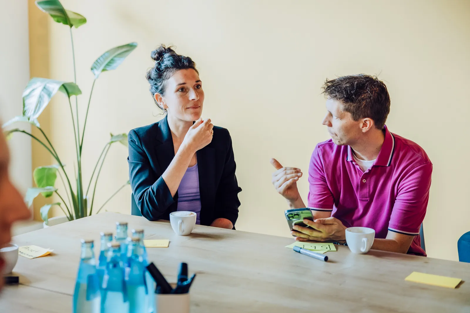 Two people sitting at a table having a discussion, with coffee cups and sticky notes in front of them. A plant is visible in the background.