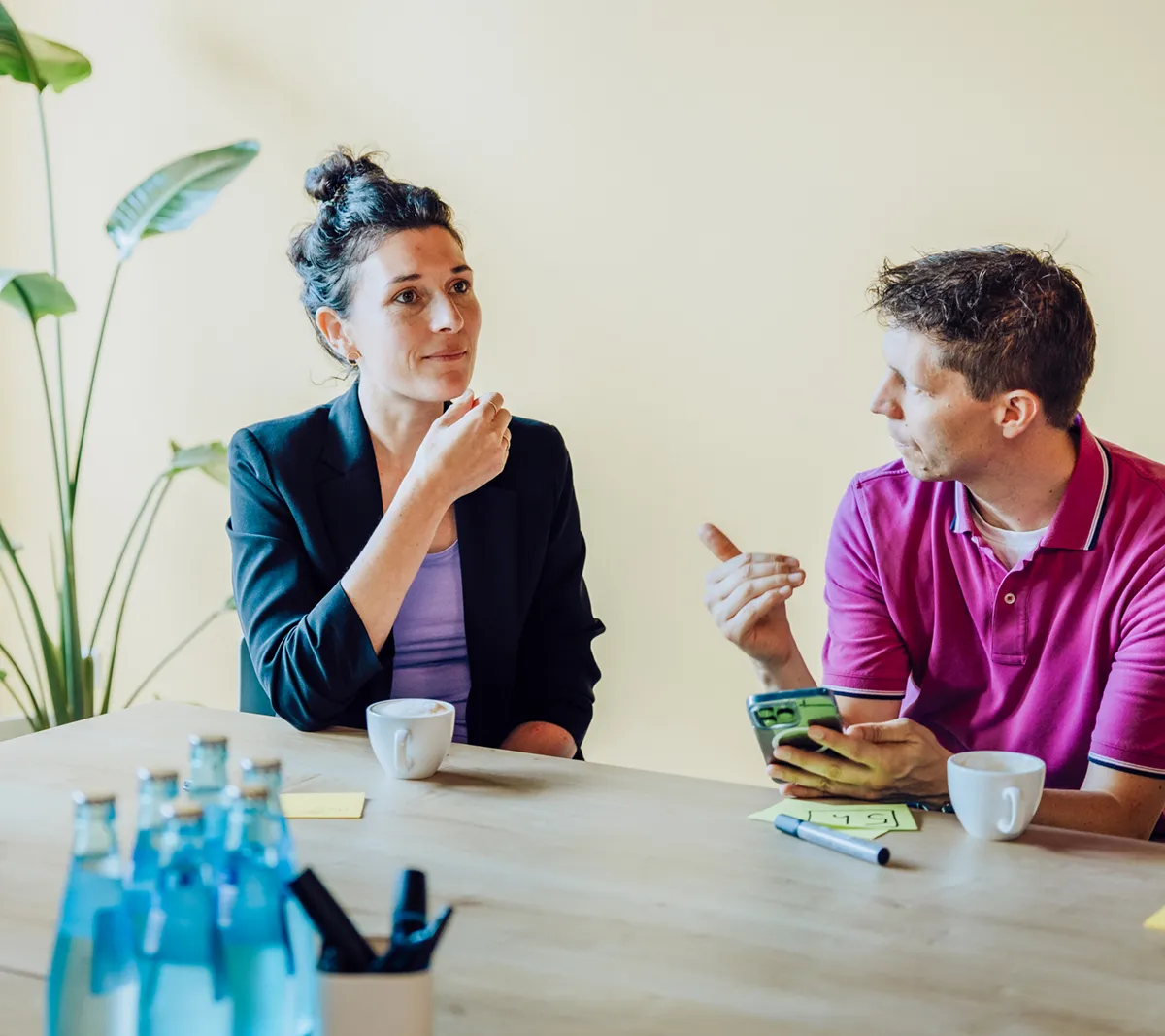 Two people sitting at a table having a discussion, with coffee cups and sticky notes in front of them. A plant is visible in the background.