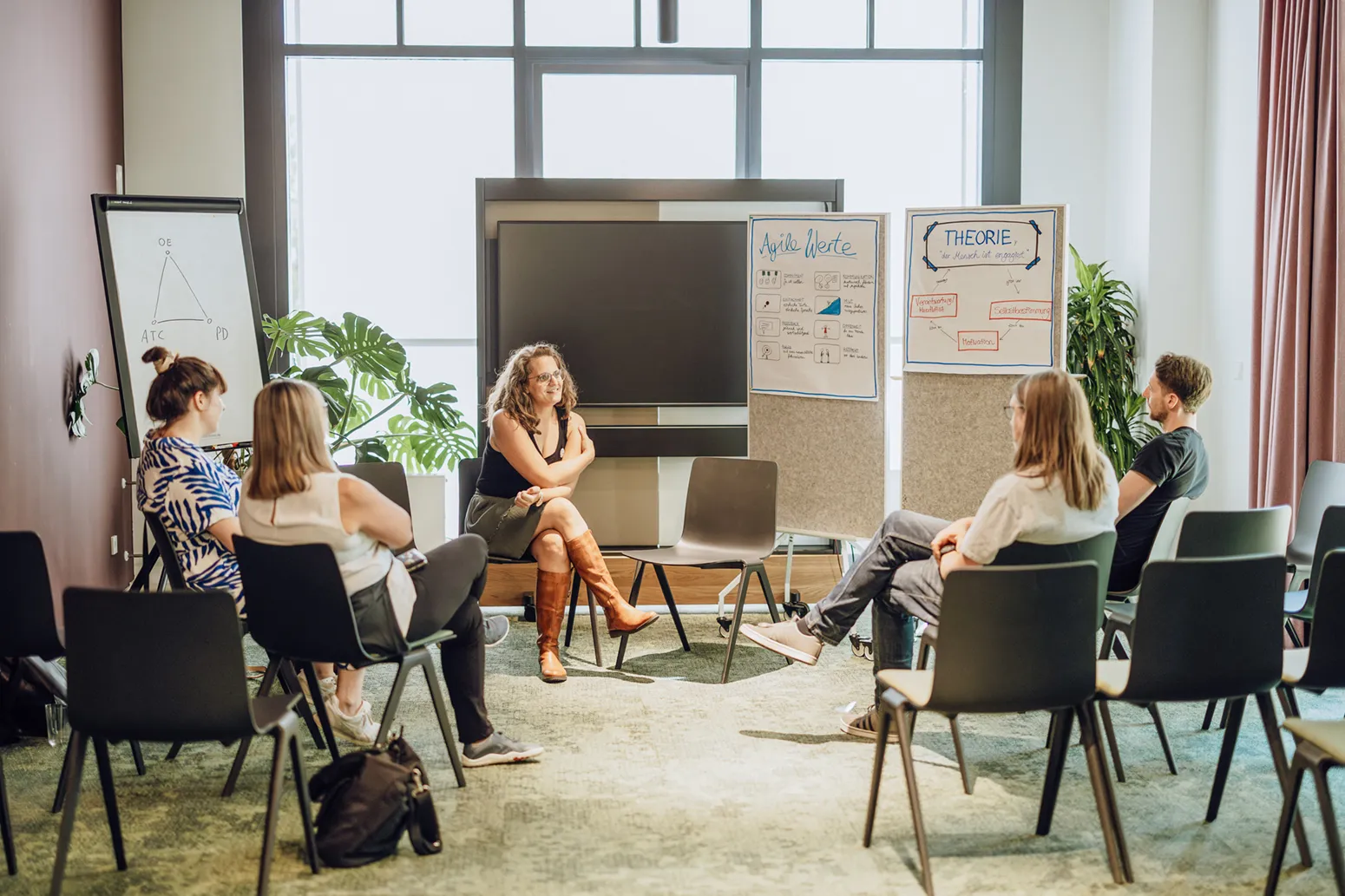 A group of people sitting in a circle in a bright room, engaged in a discussion. There are flipcharts with diagrams and notes in the background.