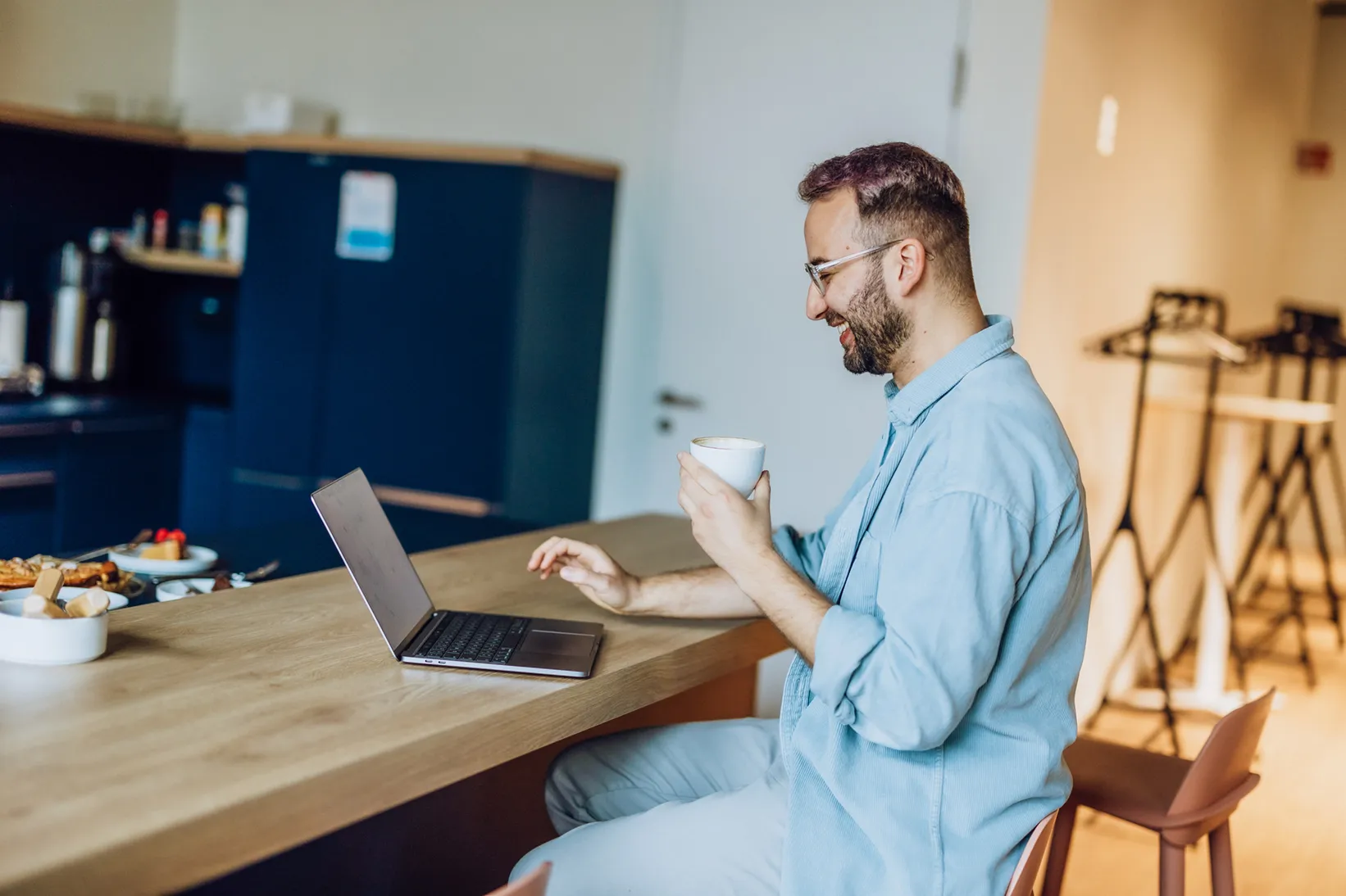 Man sitting at a kitchen counter with a laptop and holding a coffee cup, smiling while looking at the screen.