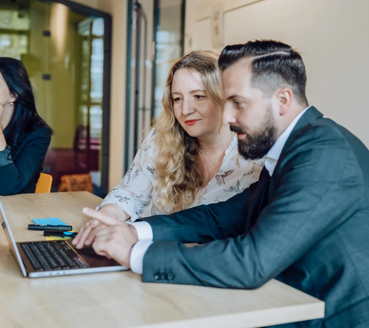 Three people in a meeting room, two focused on a laptop and one listening attentively.