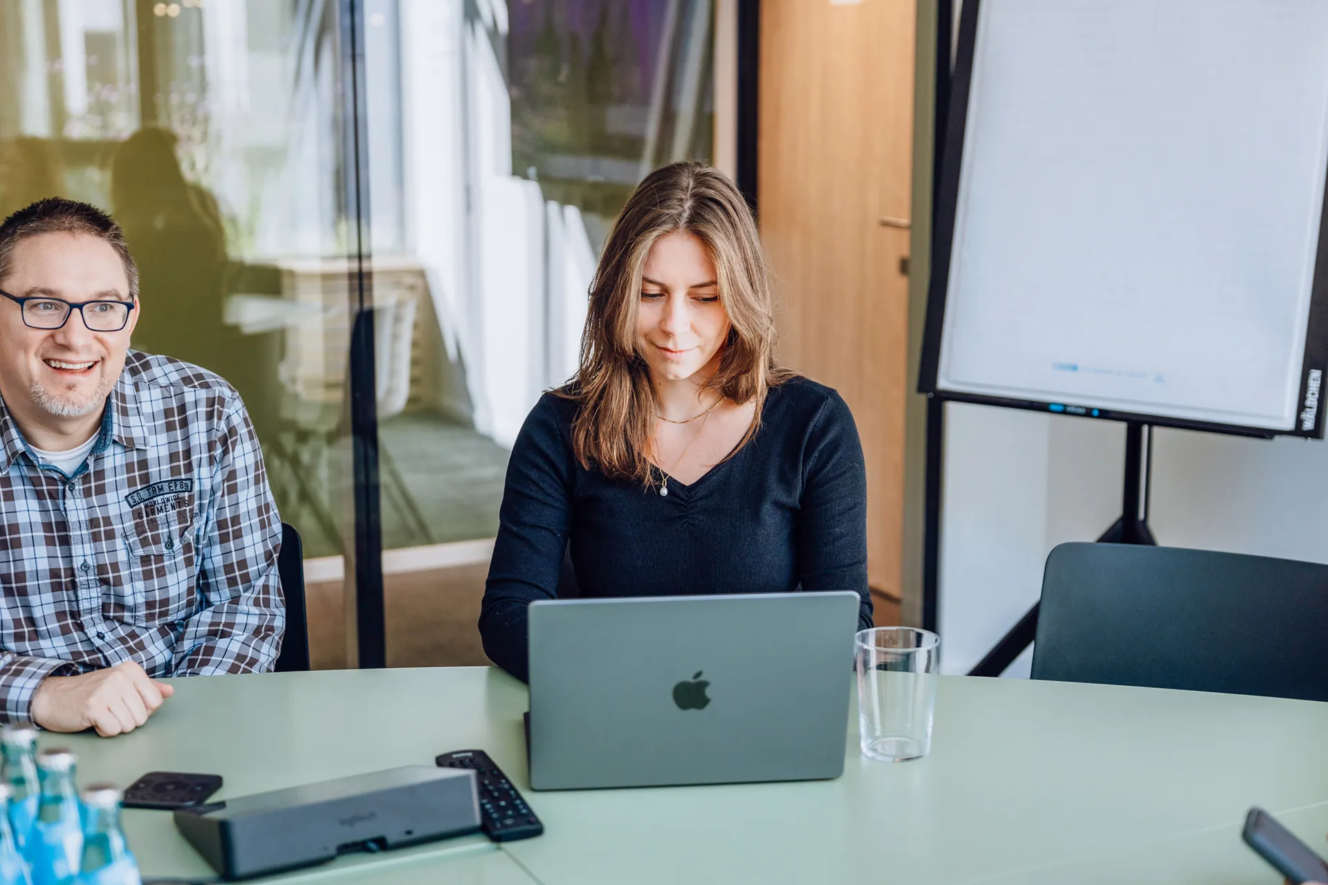 A woman working on a laptop at a green table in an office setting, with a man smiling beside her and a flipchart in the background.
