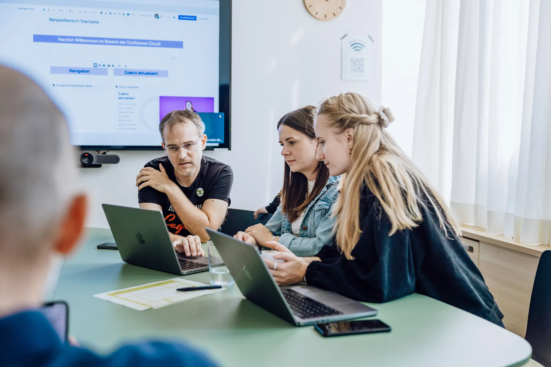 A group of four people sitting around a table with laptops, engaged in a discussion. A large screen in the background displays a webpage. The room is well-lit with natural light from a window.