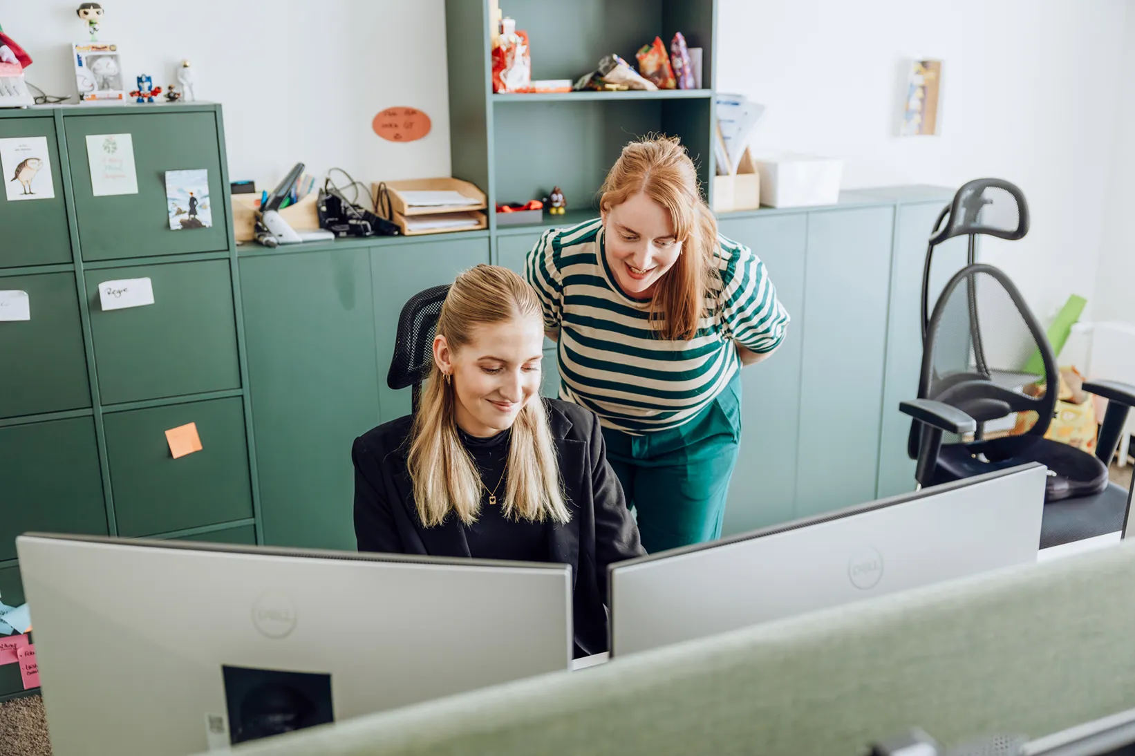 Two women in an office setting, one sitting at a desk with a computer and the other standing beside her, both smiling and looking at the screen.