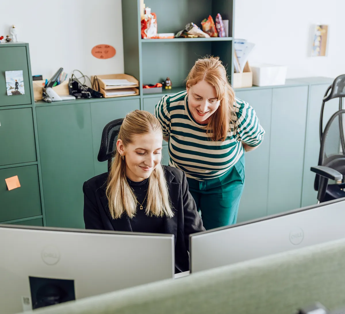 Two women in an office setting, one sitting at a desk with a computer and the other standing beside her, both smiling and looking at the screen.